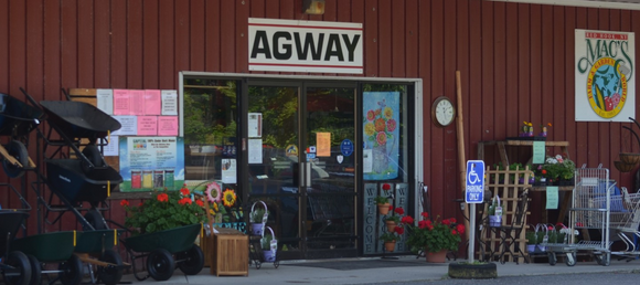 The red storefront of Mac's Agway. On the left side of the door, there are wheelbarrows, one of which contains flowers. On the right, there are plants and flowers in pots.